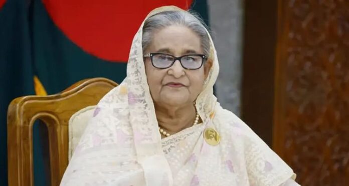 সাবেক স্বৈরাচার প্রধানমন্ত্রী শেখ হাসিনা চশমা পরে সাদা রঙের শাড়ি বা ওড়না পরিহিত অবস্থায় বসে আছেন। পেছনে বাংলাদেশের পতাকা দেখা যাচ্ছে।